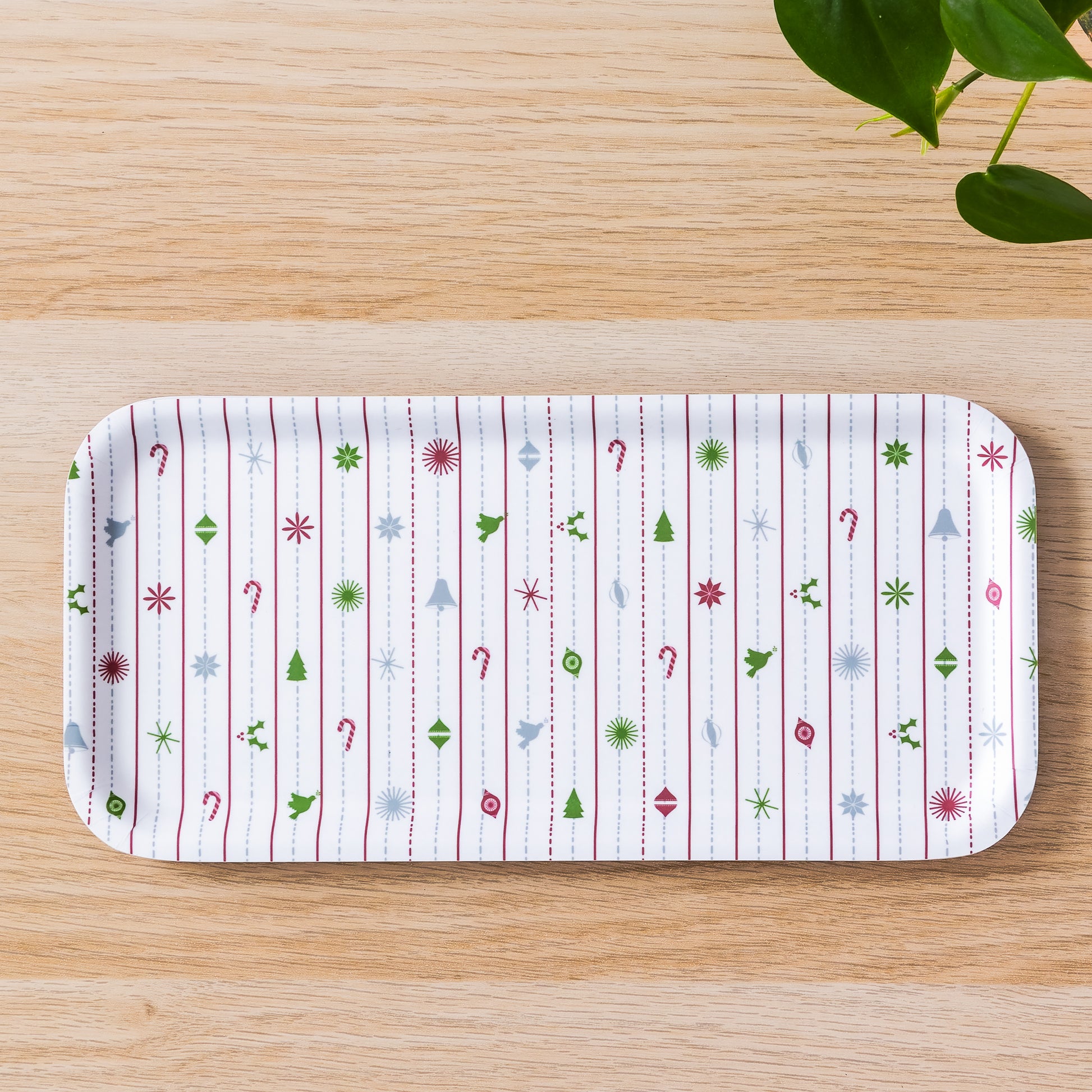 Festive birch tray with modern Christmas pattern featuring colorful ornaments, trees, candy canes, and snowflakes on white background, displayed on wooden surface with green plant leaves visible