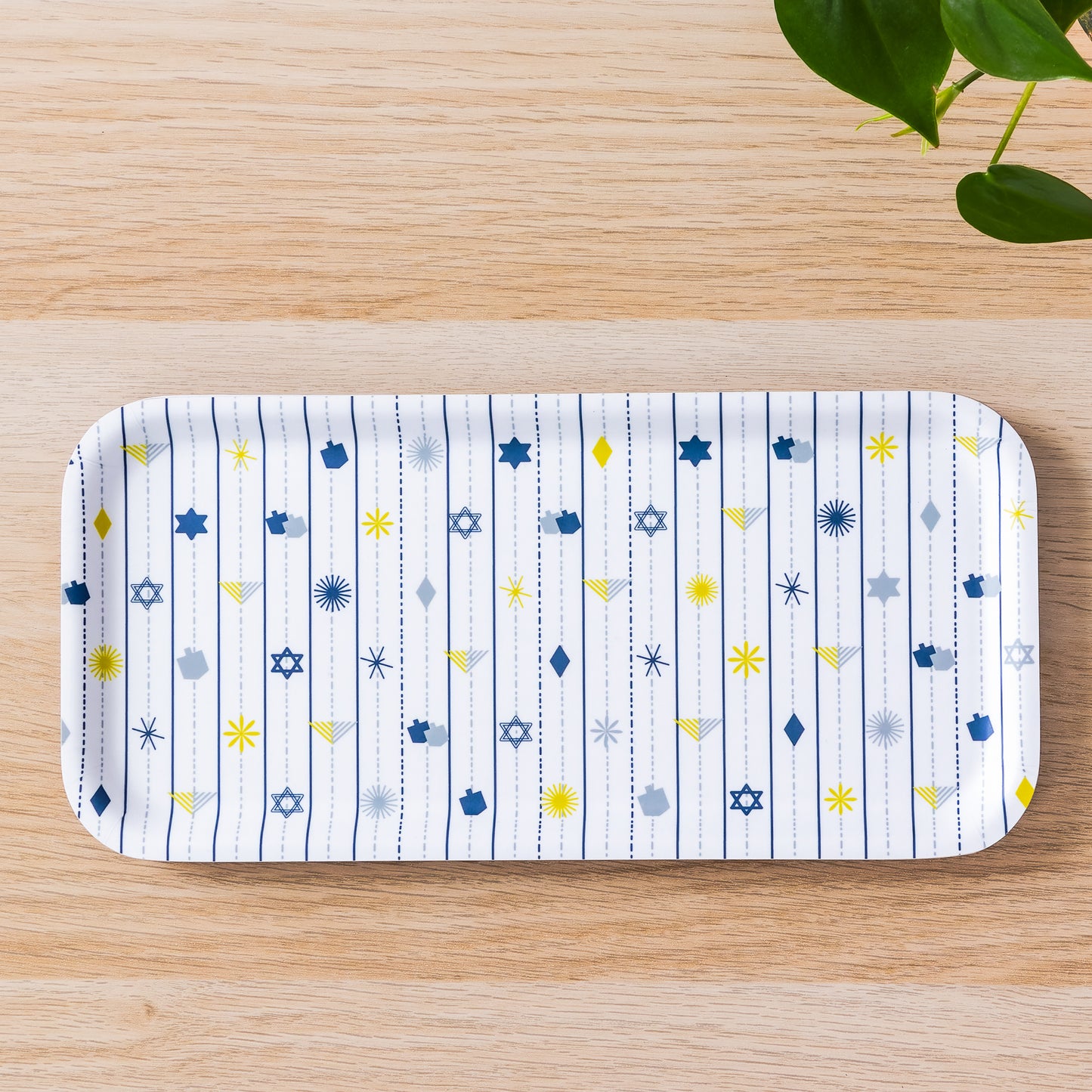 Hanukkah birch tray with white background and festive pattern of blue and yellow Stars of David, dreidels, and menorah symbols arranged in vertical stripes, displayed on wooden surface with green plant leaves