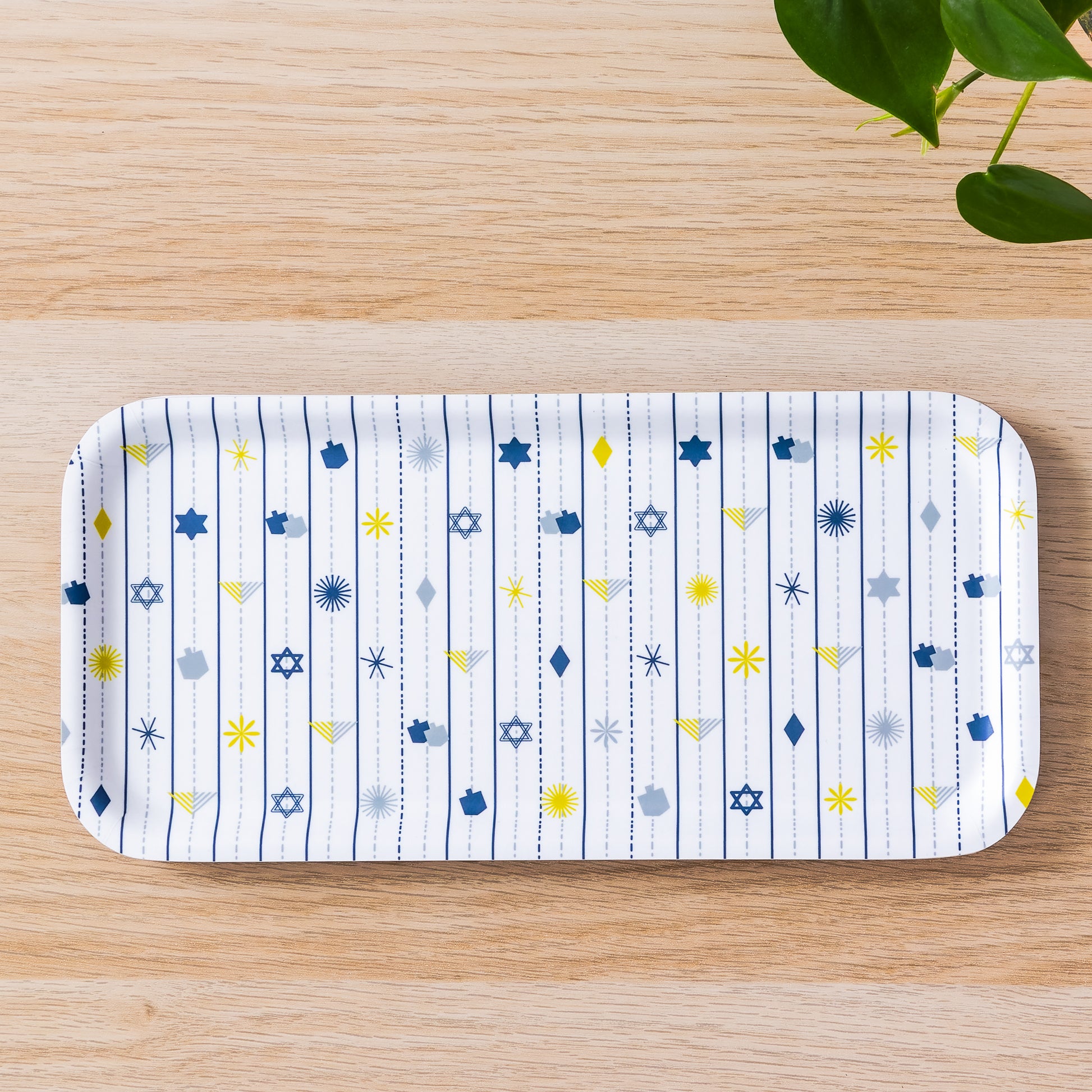 Hanukkah birch tray with white background and festive pattern of blue and yellow Stars of David, dreidels, and menorah symbols arranged in vertical stripes, displayed on wooden surface with green plant leaves