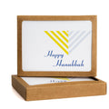 Modern Hanukkah card with stylized menorah design in yellow and gray stripes, blue "Happy Hanukkah" text, displayed in kraft paper packaging box. Eco-friendly A2 size card for festive holiday greetings.
